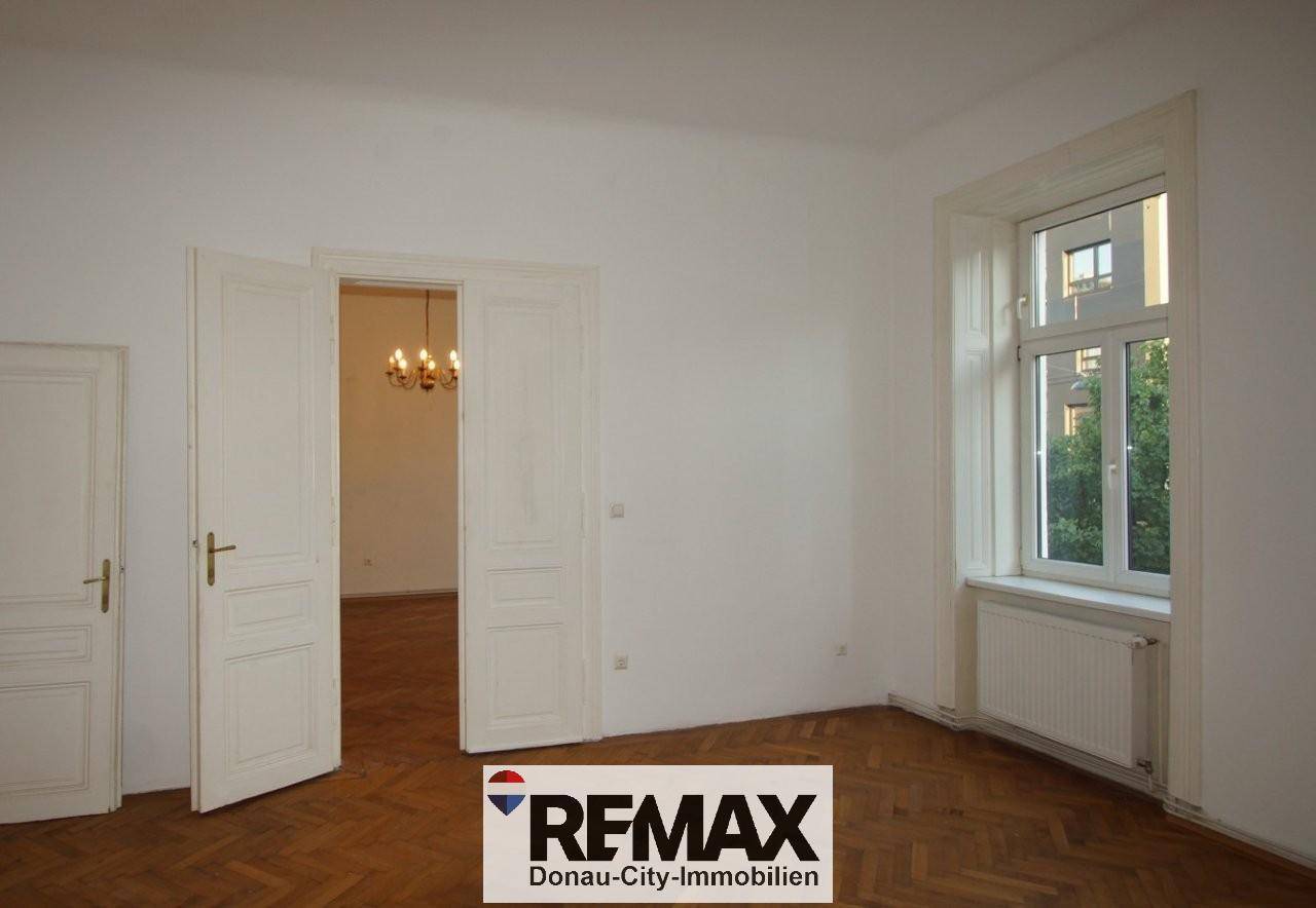 Helles Zimmer mit Fischgrätparkett und einem Fenster, das zu einem weiteren Raum führt.