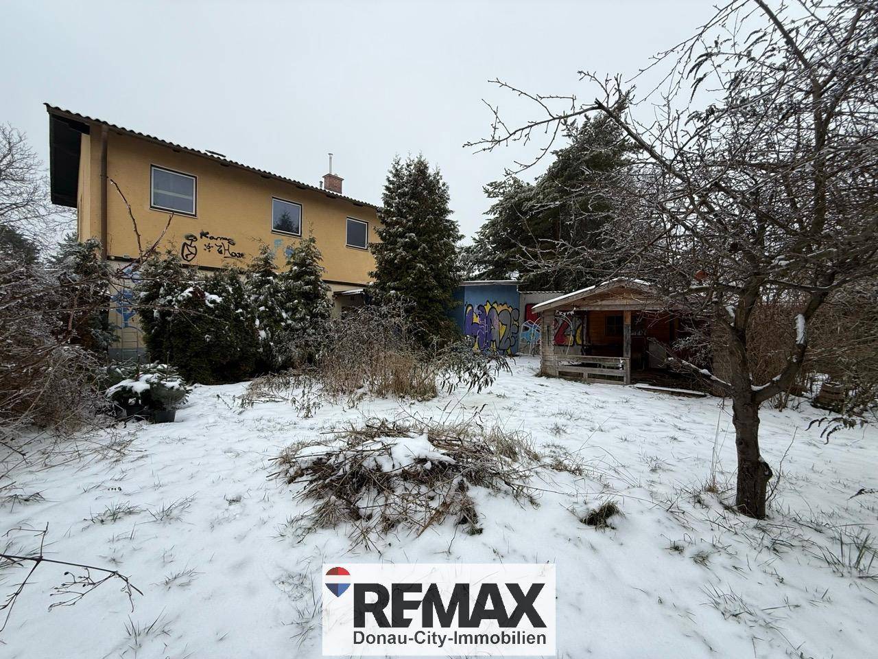 Weitläufiger Garten im Winter mit Schnee und dem Haus im Hintergrund, umgeben von Bäumen.