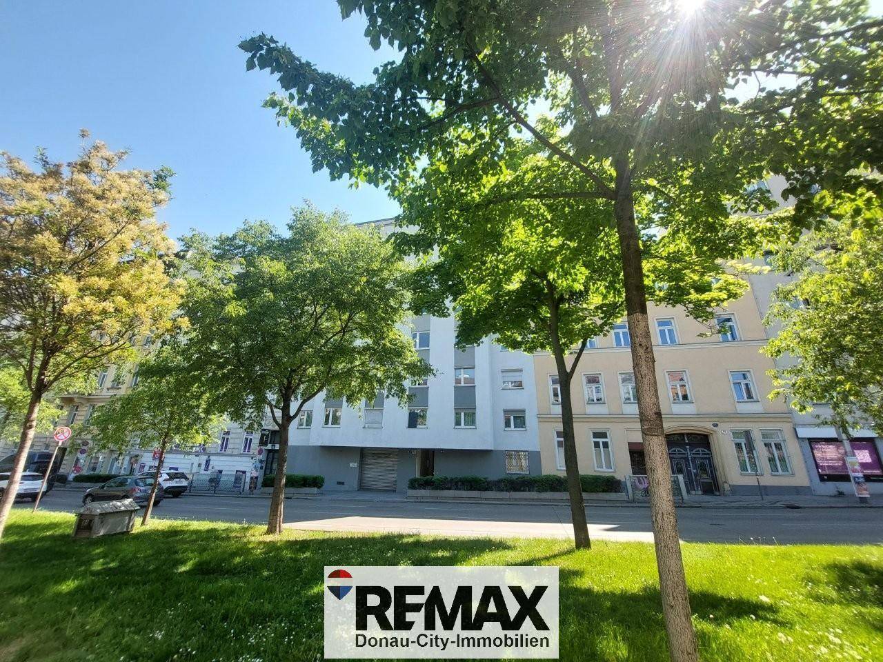 Straßenansicht eines modernen Wohngebäudes mit Bäumen und viel Grün in sonniger Umgebung.