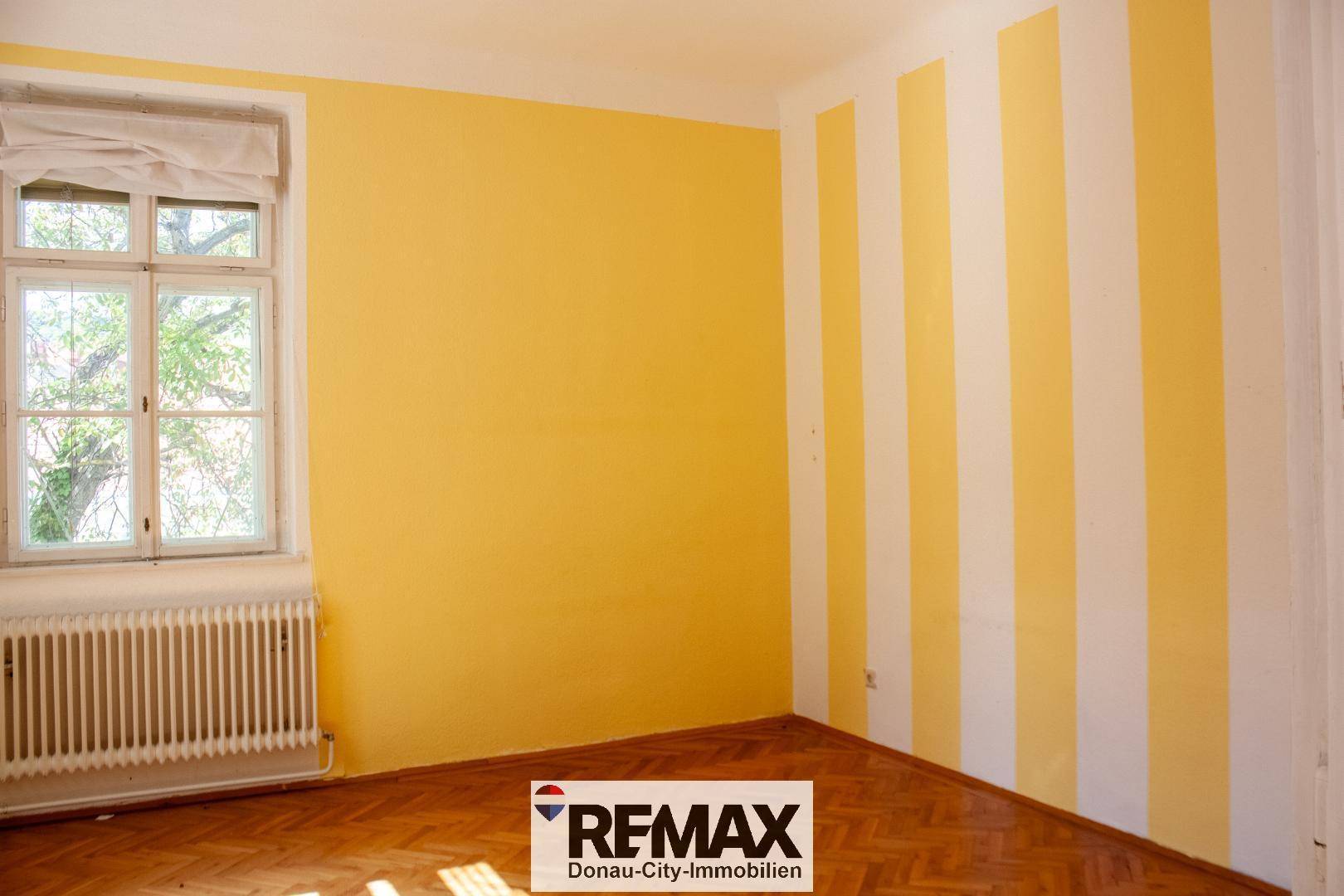 Helles Zimmer mit gelb-weiß gestreiften Wänden, Holzparkettboden und einem Fenster mit Heizkörper.