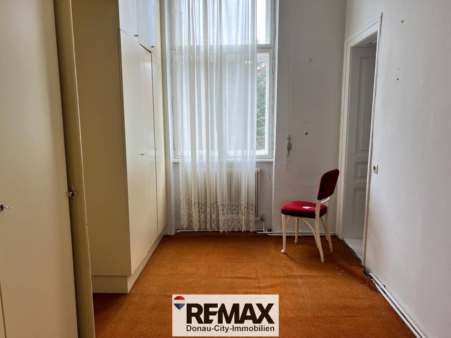 Schlafzimmer mit orangefarbenem Teppichboden, praktischen Einbauschränken und einem Fenster mit hellen Vorhängen.