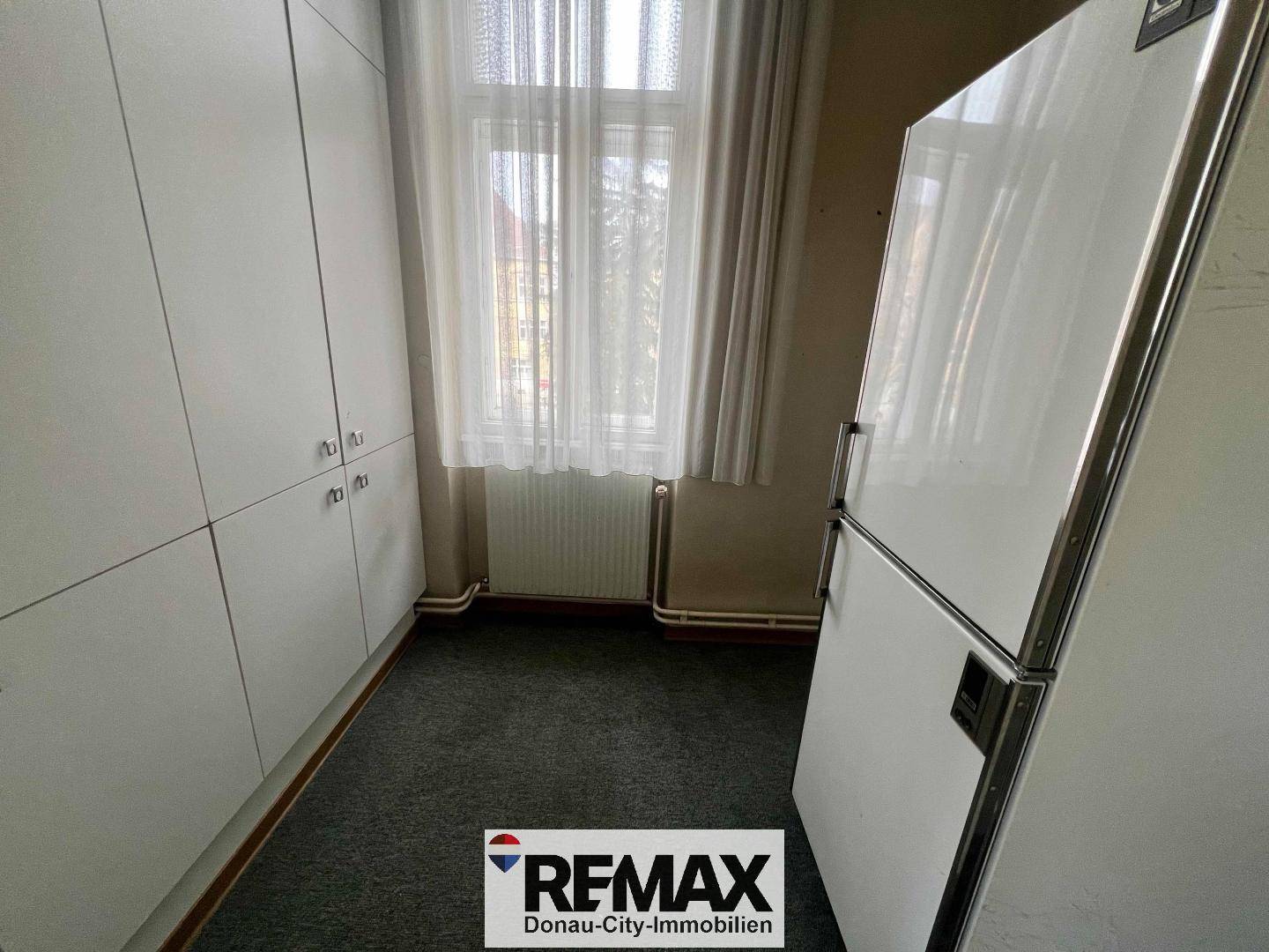 Kleiner Raum mit Teppichboden, Fenster und einem Kühlschrank.