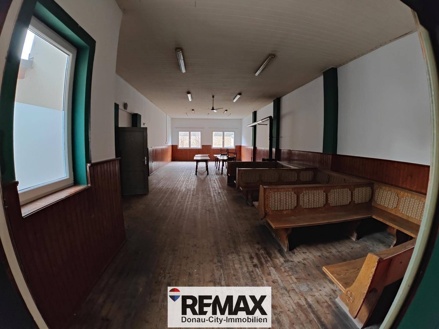 Großer Saal mit Holzboden, langen Sitzbänken und mehreren Fenstern für Tageslicht.