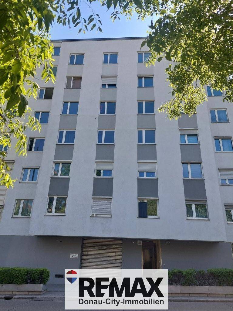 Moderne Fassade eines Mehrfamilienhauses mit vielen Fenstern und Rollläden, umgeben von Bäumen.