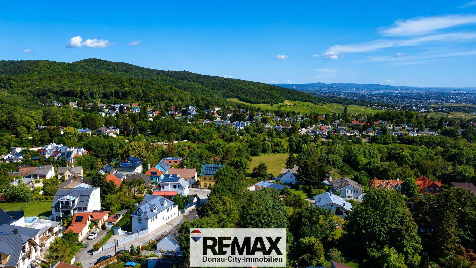 Weitläufige Luftaufnahme einer Wohnsiedlung eingebettet in eine grüne Hügellandschaft unter blauem Himmel.