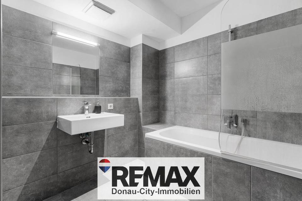 Modernes Badezimmer mit grauen Fliesen, wandhängendem Waschbecken und Badewanne mit Duschwand.