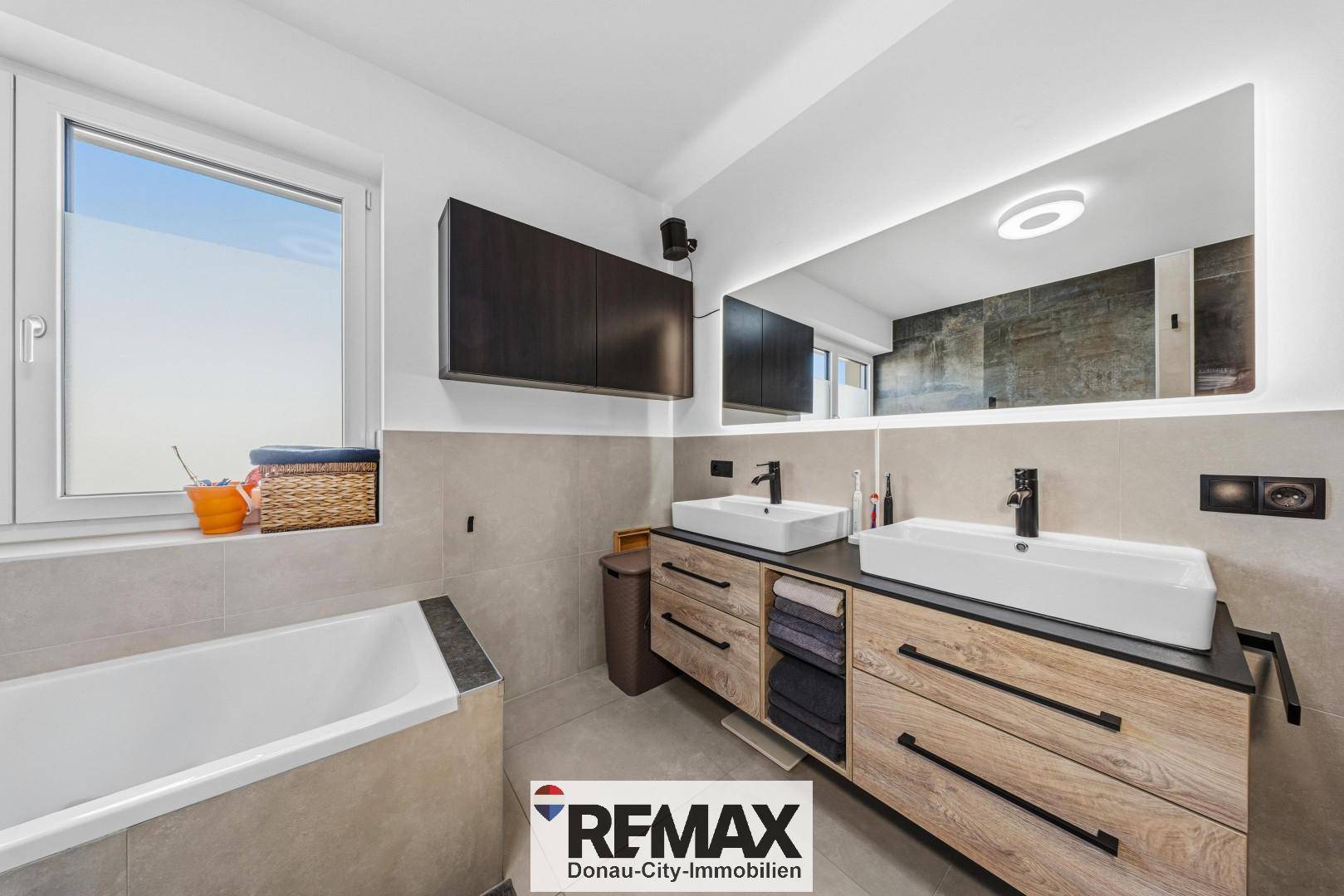 Modernes Badezimmer mit Doppelwaschbecken, beleuchtetem Spiegel und Badewanne unter dem Fenster.