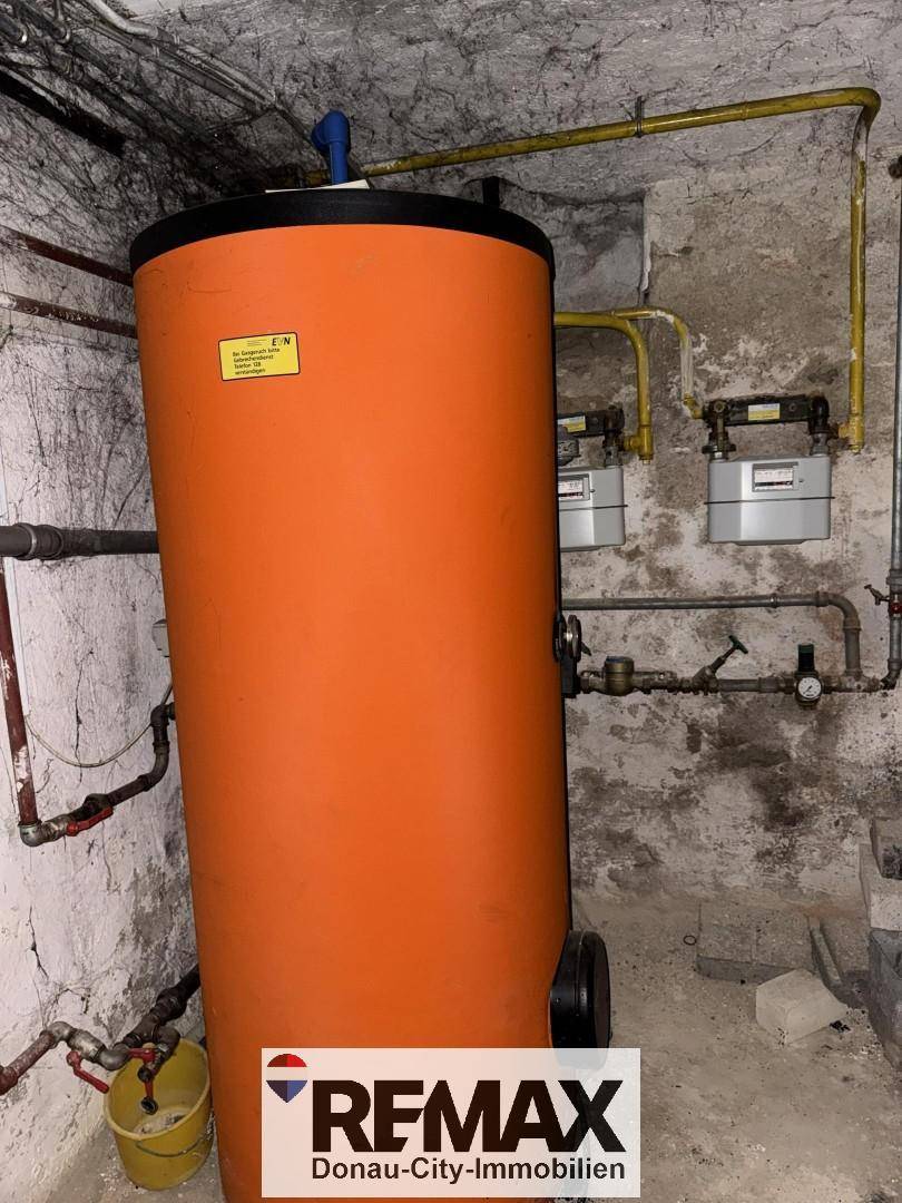 Kellerraum mit einem großen orangefarbenen Warmwasserspeicher und sichtbaren Rohrleitungen an den Wänden.