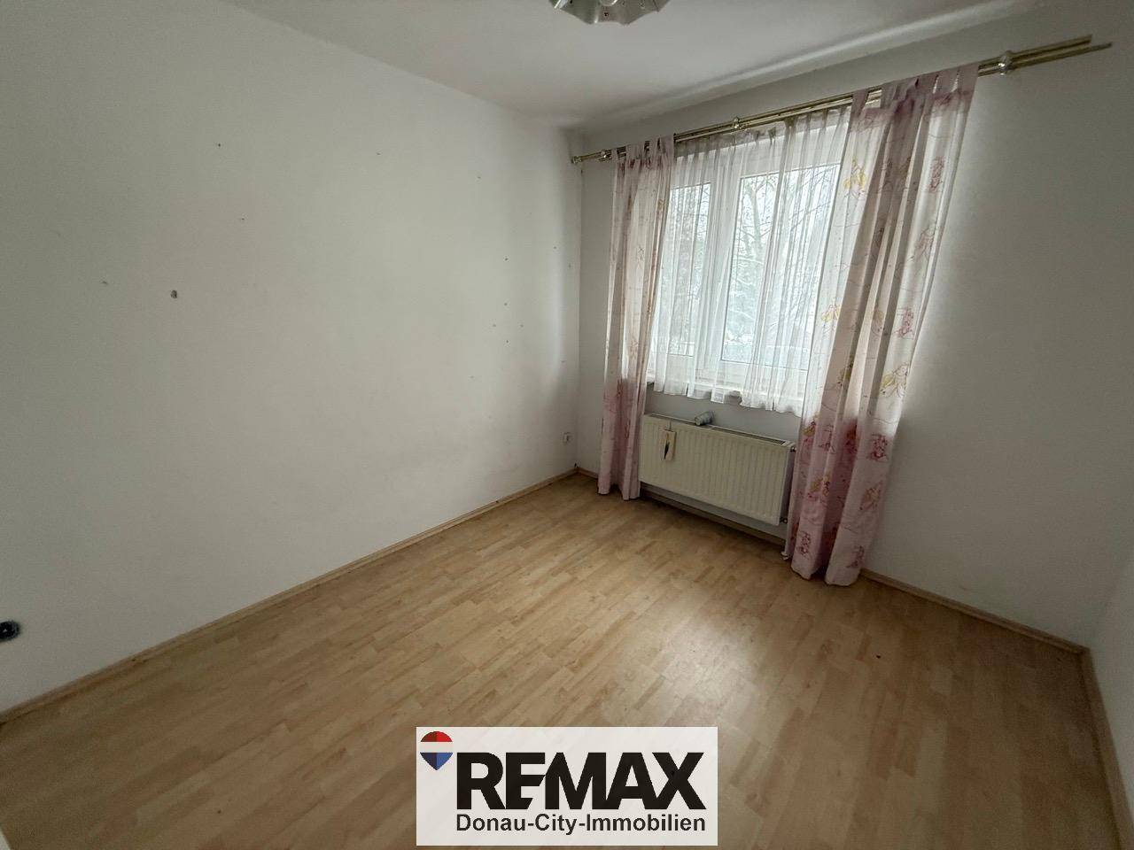Schlafzimmer mit Laminatboden und Vorhängen am Fenster, das auf die Umgebung blickt.