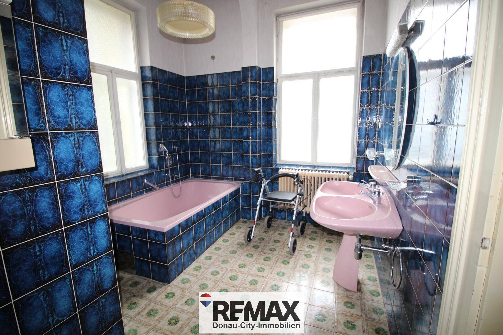 Badezimmer im 80er-Jahre-Stil mit blauen Fliesen, rosa Badewanne und Doppelwaschbecken.
