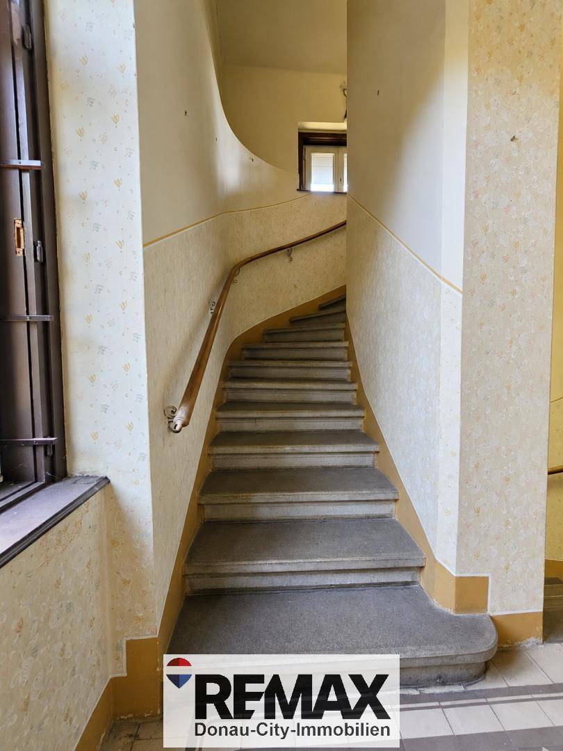 Treppenhaus mit geschwungener Treppe, Tapeten und Holzgeländer, das Renovierungsbedarf zeigt.