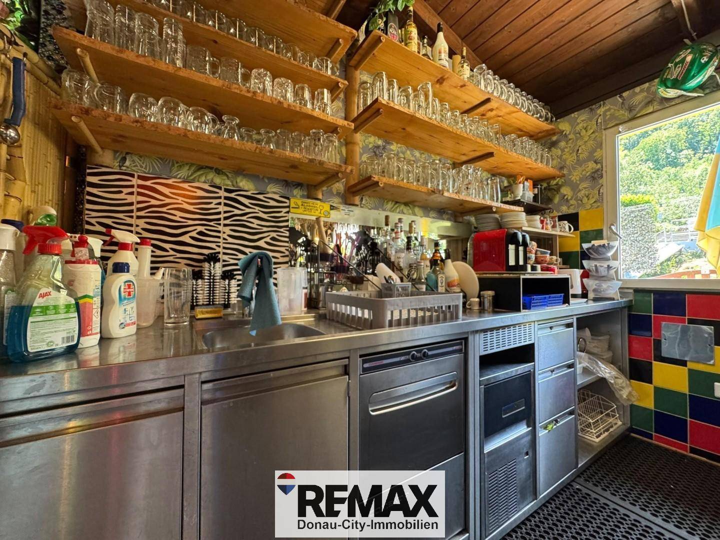 Küchenbereich hinter der Bar mit Spüle, offenen Holzregalen voller Gläser und Arbeitsflächen aus Edelstahl.