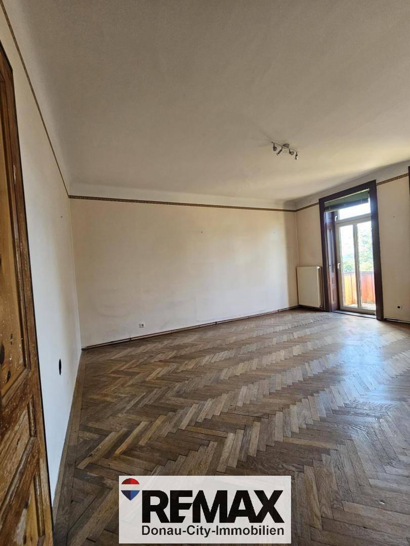 Leerer Raum mit Fischgrätparkett, hohen Decken und einem Fenster mit Blick ins Grüne.