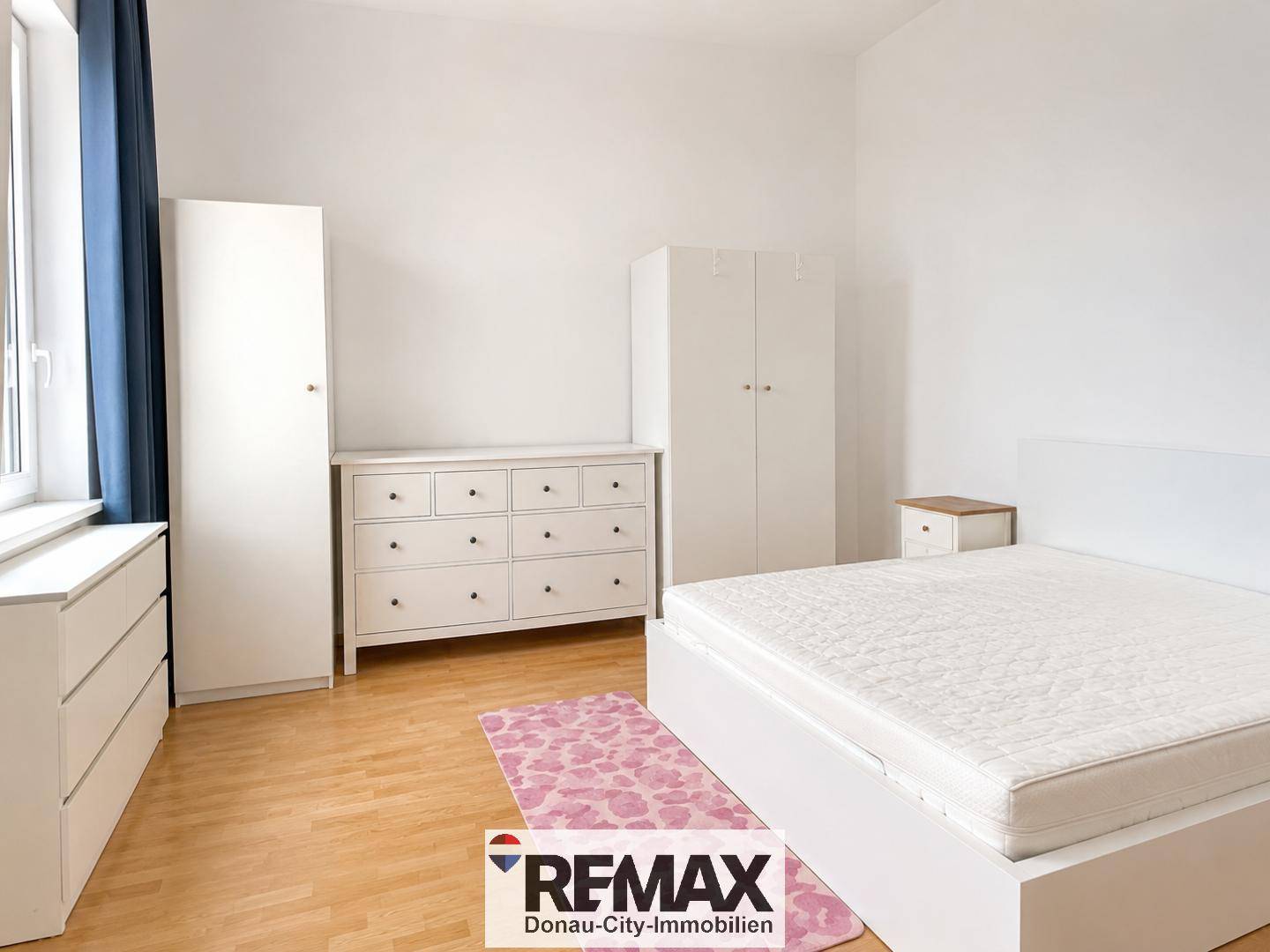 Schlafzimmer mit weißem Bett, Kommoden, Kleiderschränken und einem Fenster mit dunklen Vorhängen.