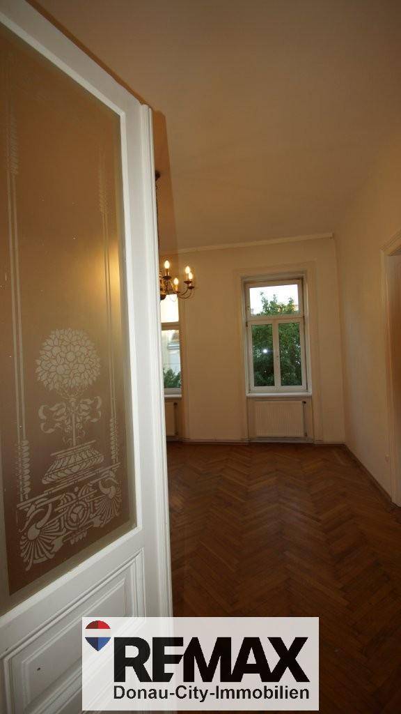 Durchblick durch eine verzierte Glastür in ein helles Zimmer mit Fischgrätparkett und Fenster.