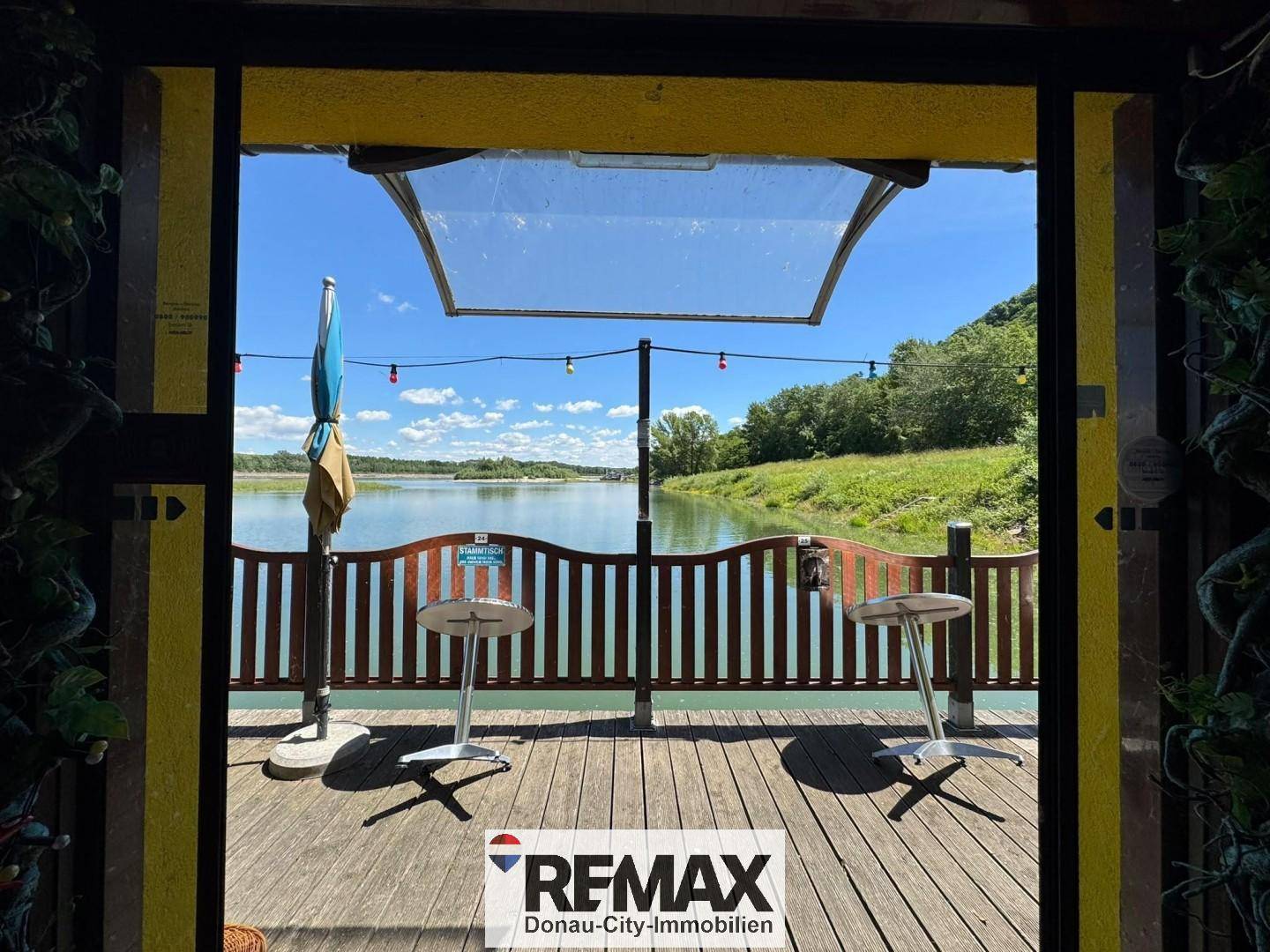 Blick von innen auf die Holzterrasse mit Tischen und Stühlen, die direkt auf den Fluss hinausgeht.