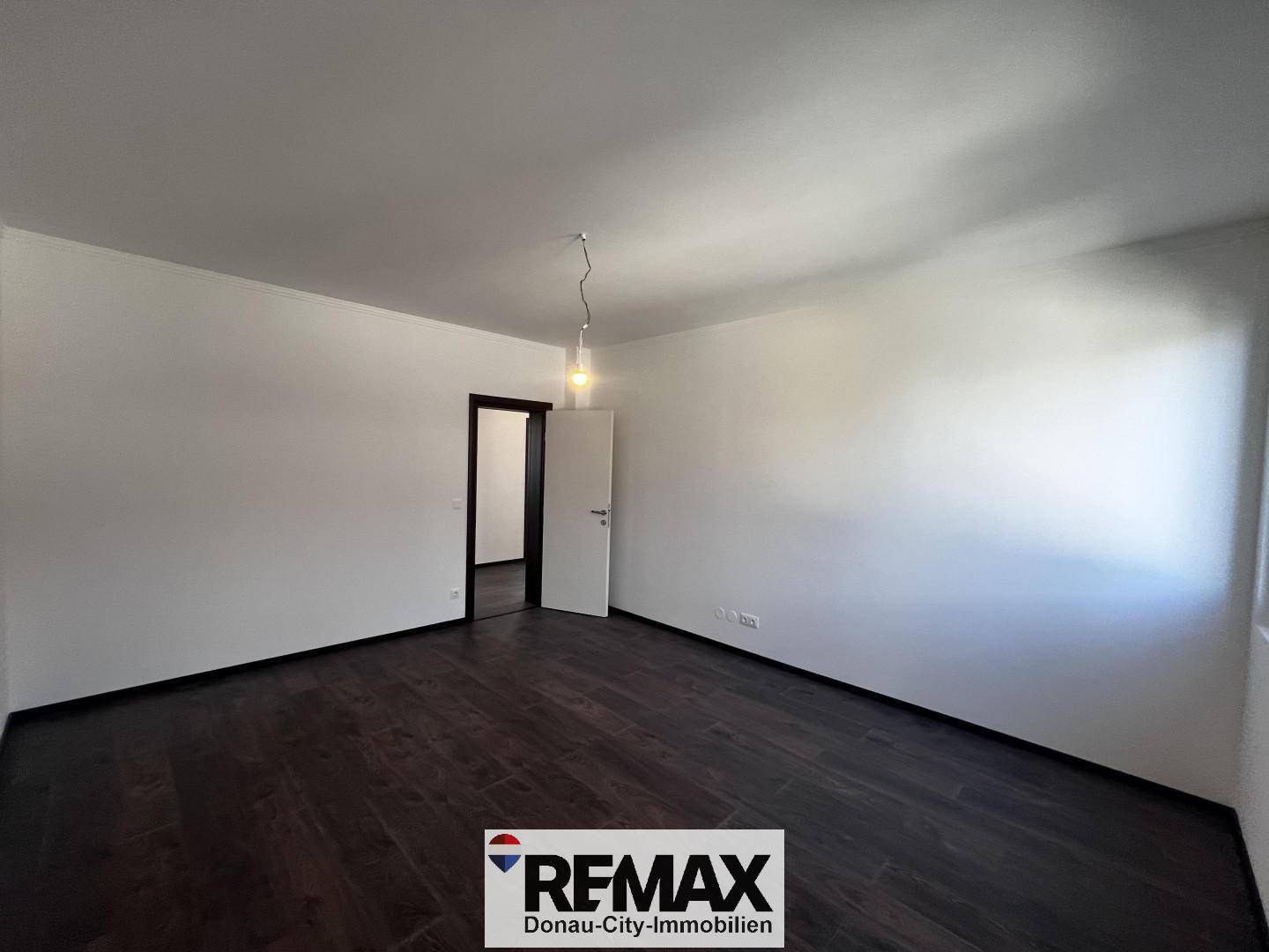 Heller Raum mit dunklem Holzboden und weiß gestrichenen Wänden, mit Zugang zu einem weiteren Zimmer.