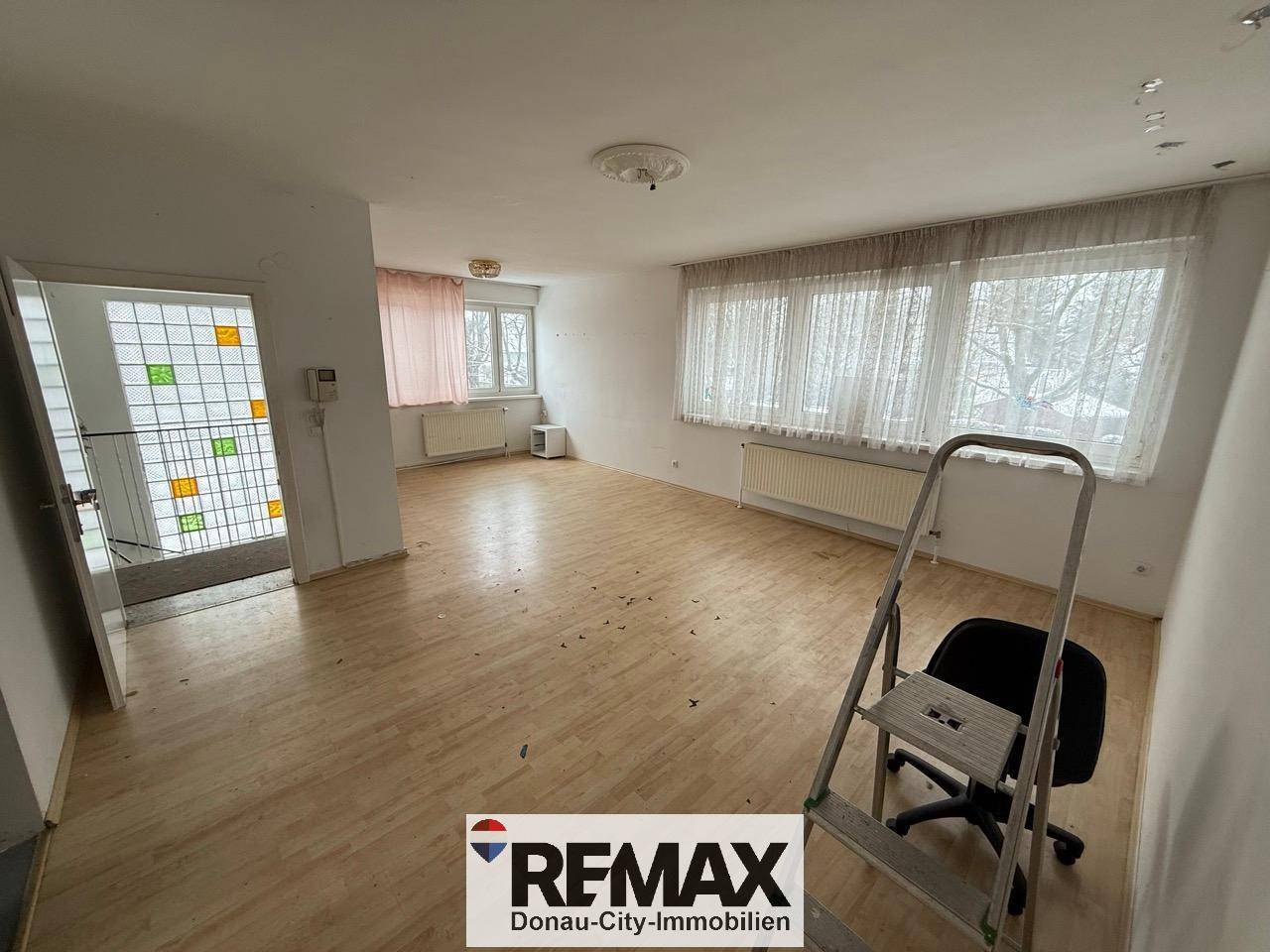 Helles, leeres Wohnzimmer mit Laminatboden und großen Fenstern, das viel Tageslicht hereinlässt.