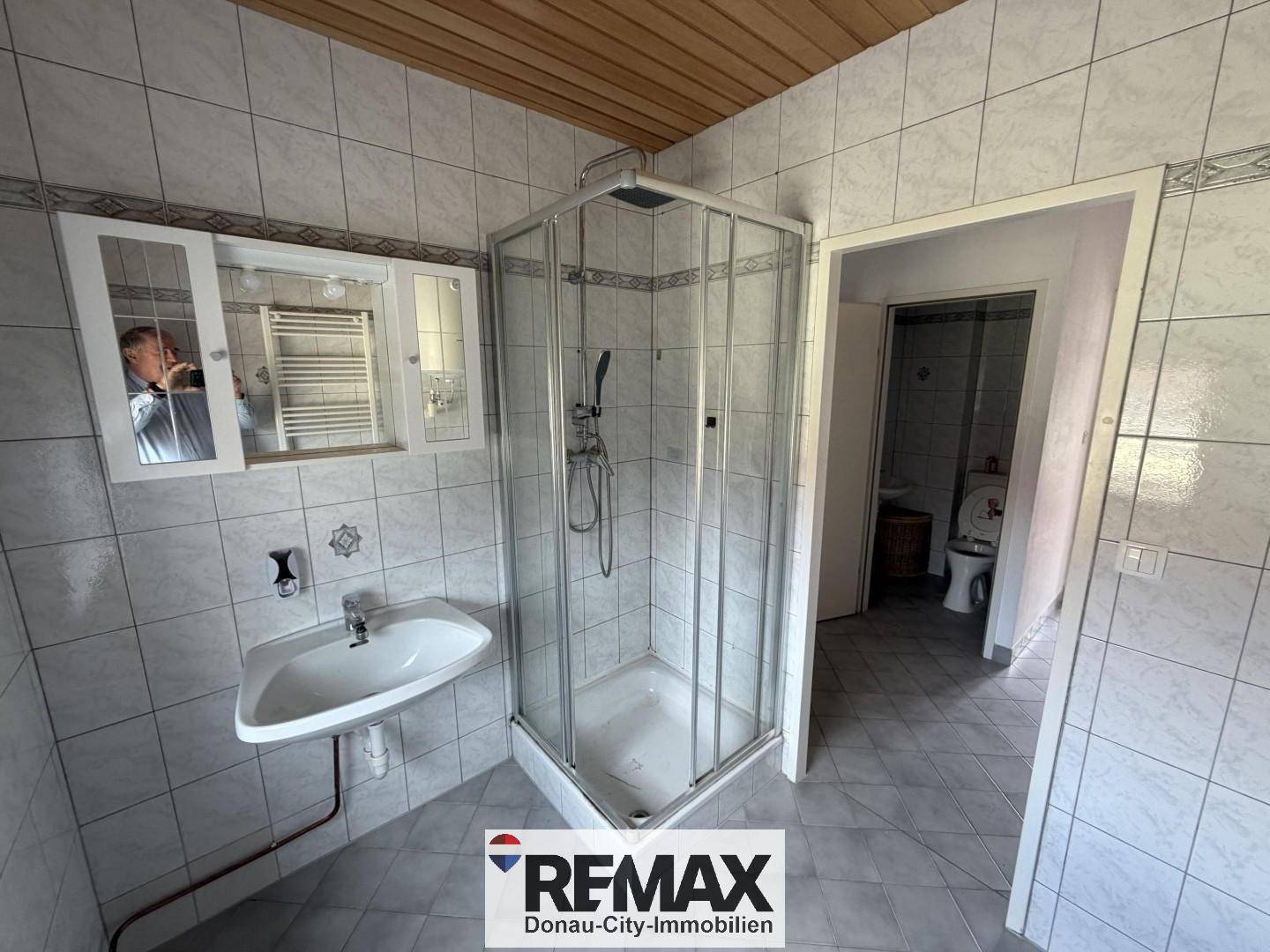 Gefliestes Badezimmer mit Duschkabine, Waschbecken und Holzdecke, angrenzend an separates WC.