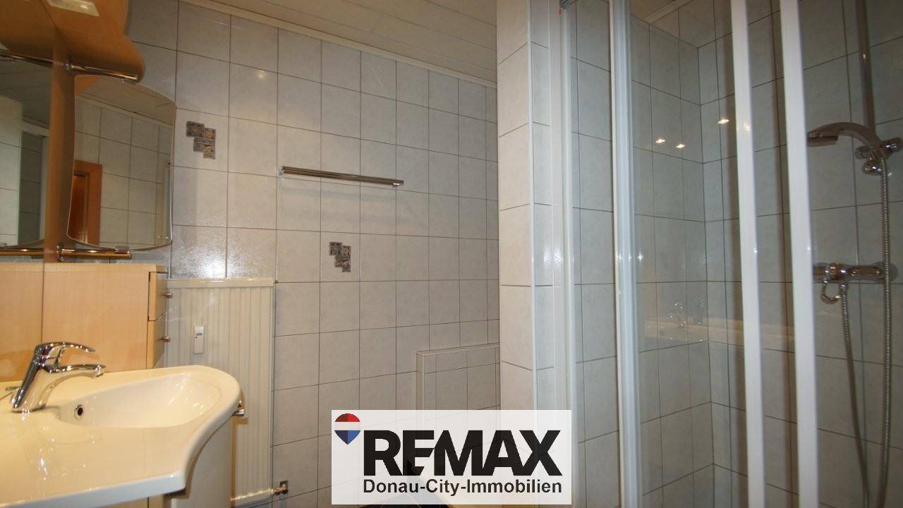 Badezimmer mit Waschbecken, Spiegel, Heizkörper und einer Duschkabine, hell gefliest.