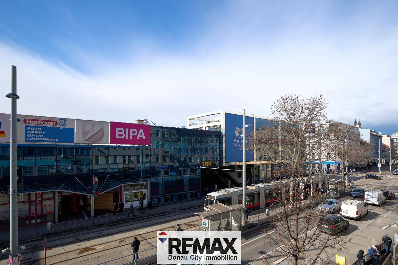 Belebtes Stadtbild mit Straßenbahn, Geschäften und Gebäuden unter blauem Himmel.