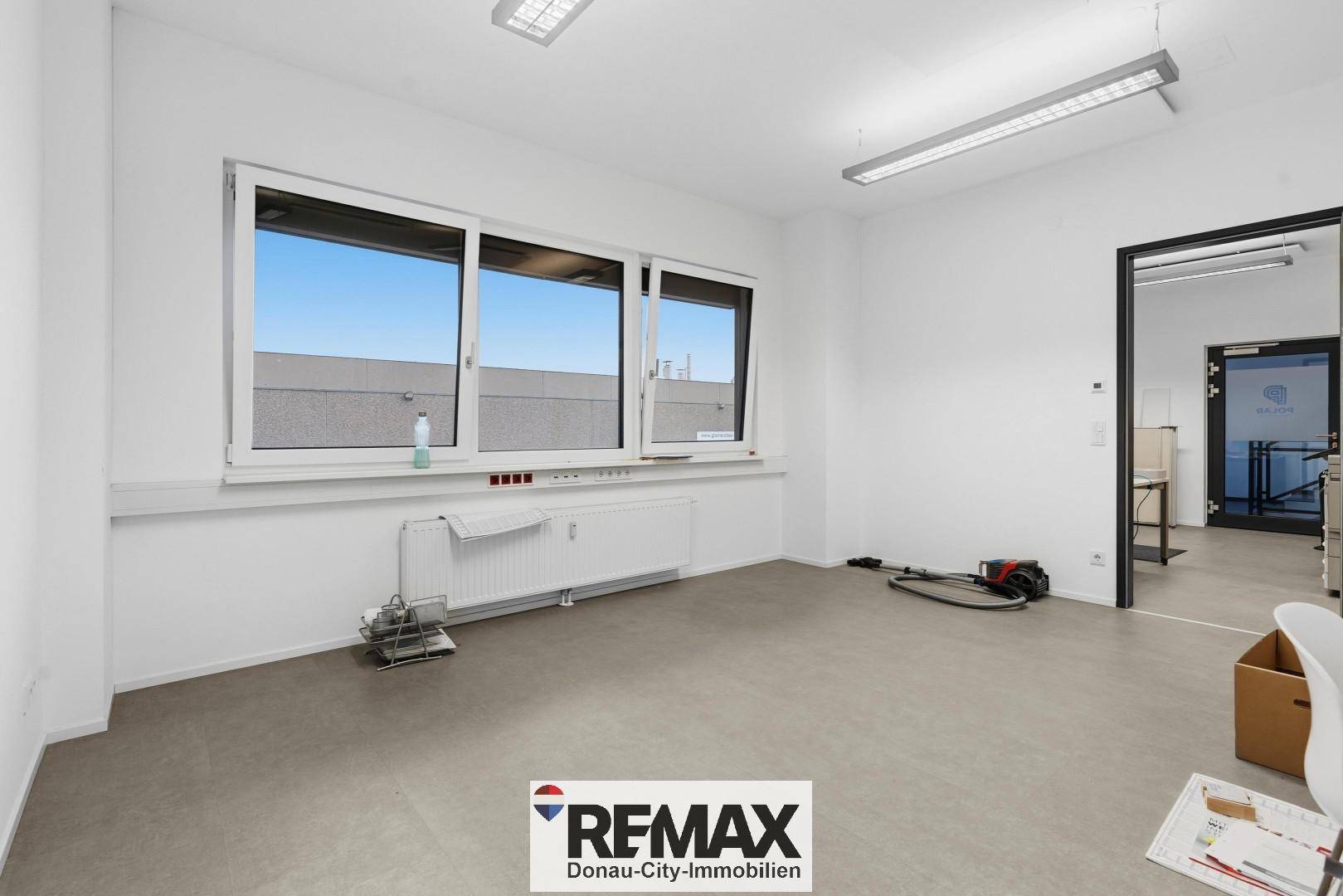Leeres Büro mit großen Fenstern, Heizkörper und Blick auf das Gebäude gegenüber.