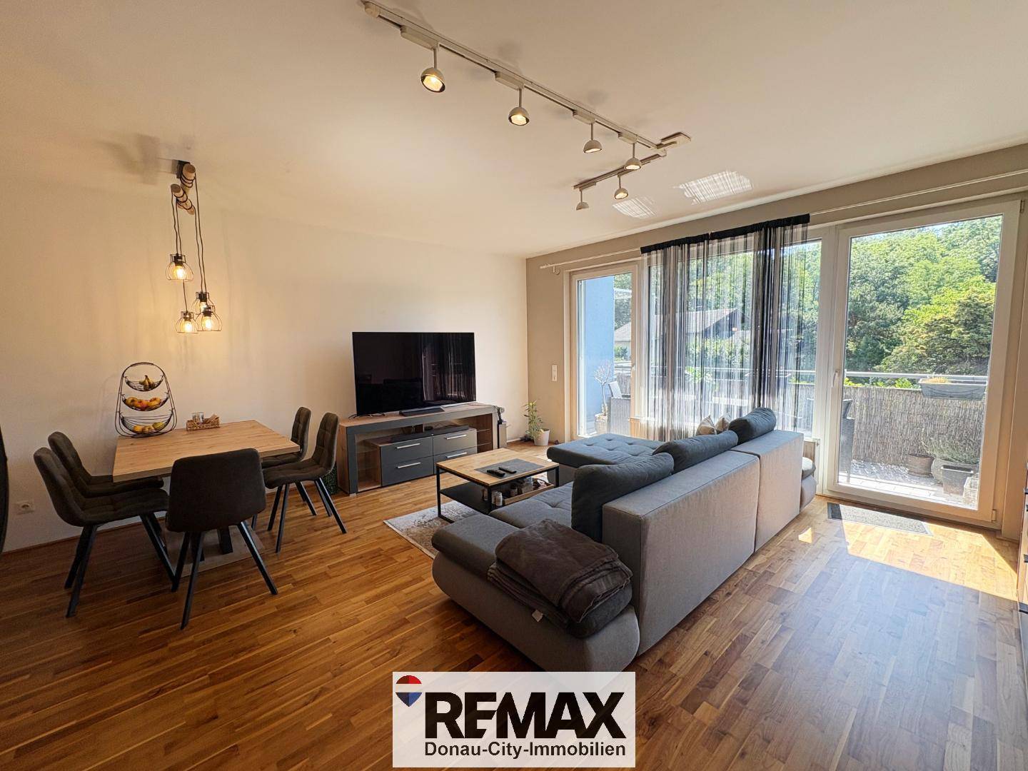 Heller Wohn- und Essbereich mit Holzboden, großem Sofa und Zugang zum Balkon.