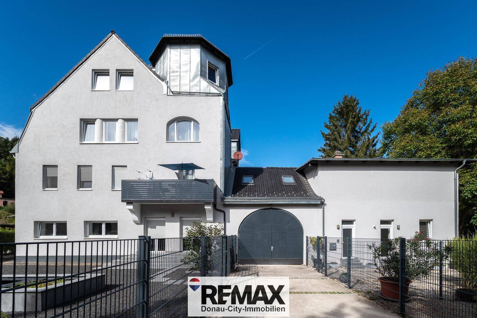 Seitenansicht des Gebäudes mit großer gewölbter Garagentür, kleinem Balkon und gepflegtem Vorgartenbereich.