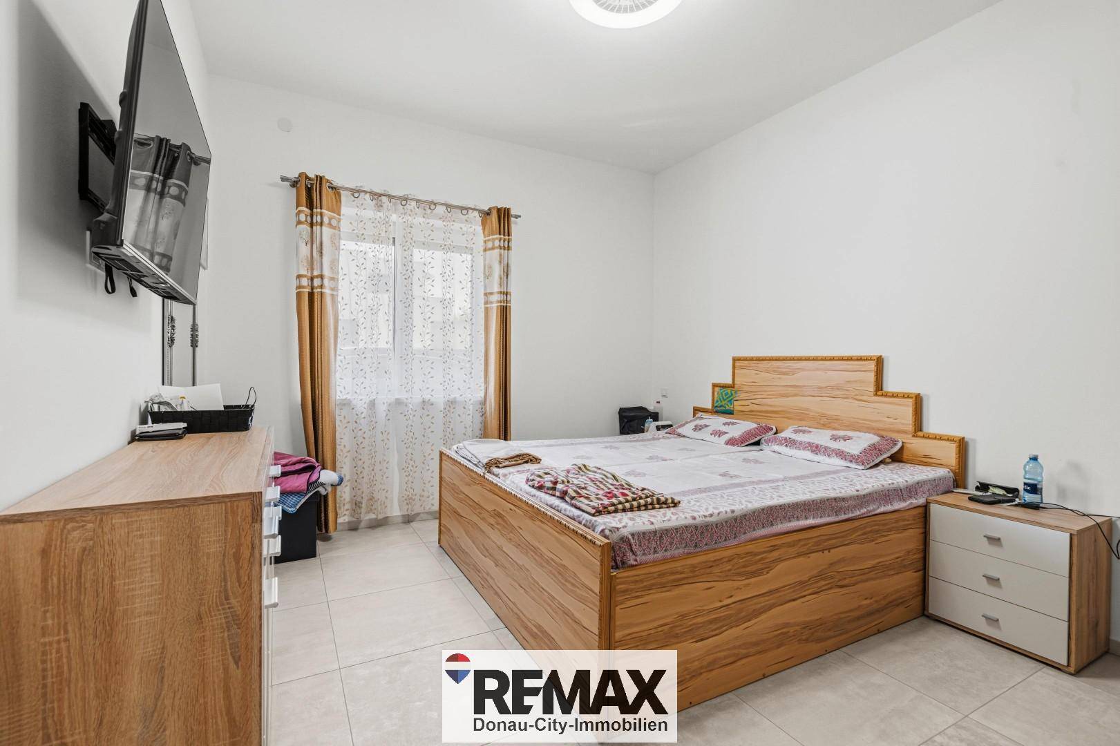 Helles Schlafzimmer mit Holzbett, Kommode und Fliesenboden, gut beleuchtet durch ein Fenster.