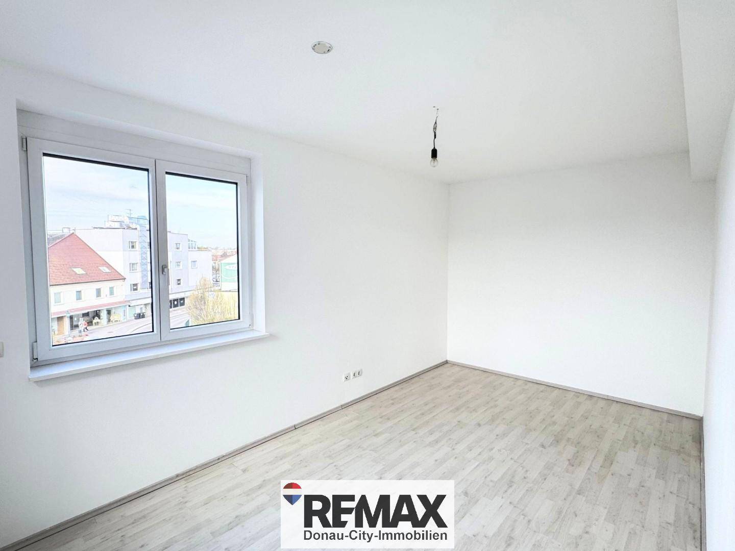 Helles Zimmer mit weiß gestrichenen Wänden, hellem Boden und Fensterblick auf die Umgebung.