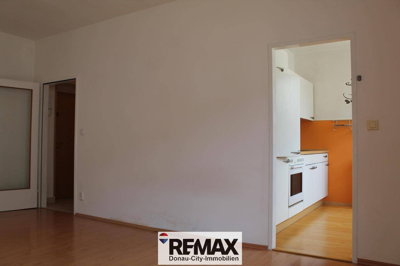 Leeres Wohnzimmer mit Laminatboden und Zugang zu einer kleinen Küche.