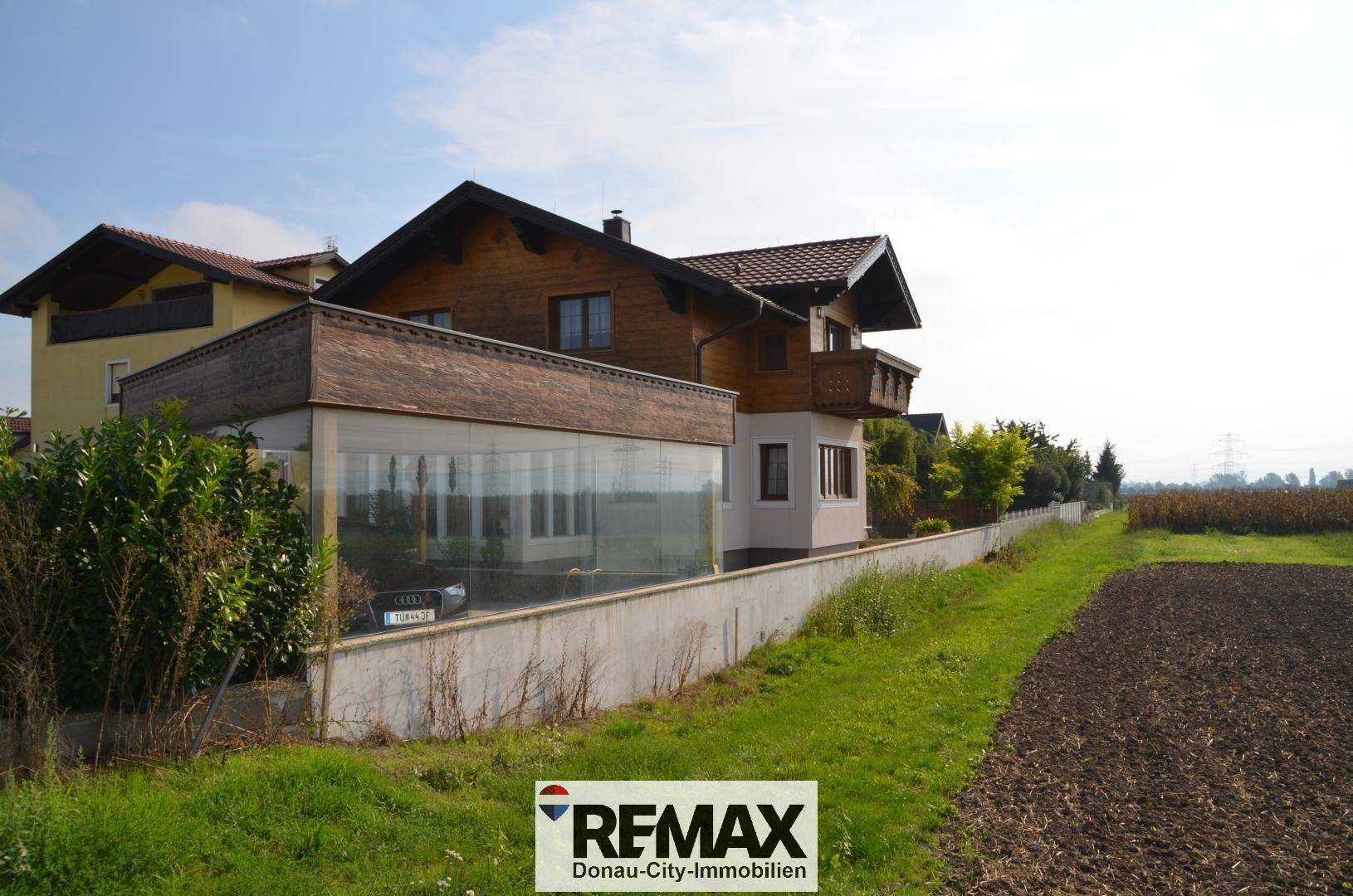 Seitenansicht des Hauses mit verglastem Anbau und angrenzendem Feld unter blauem Himmel.