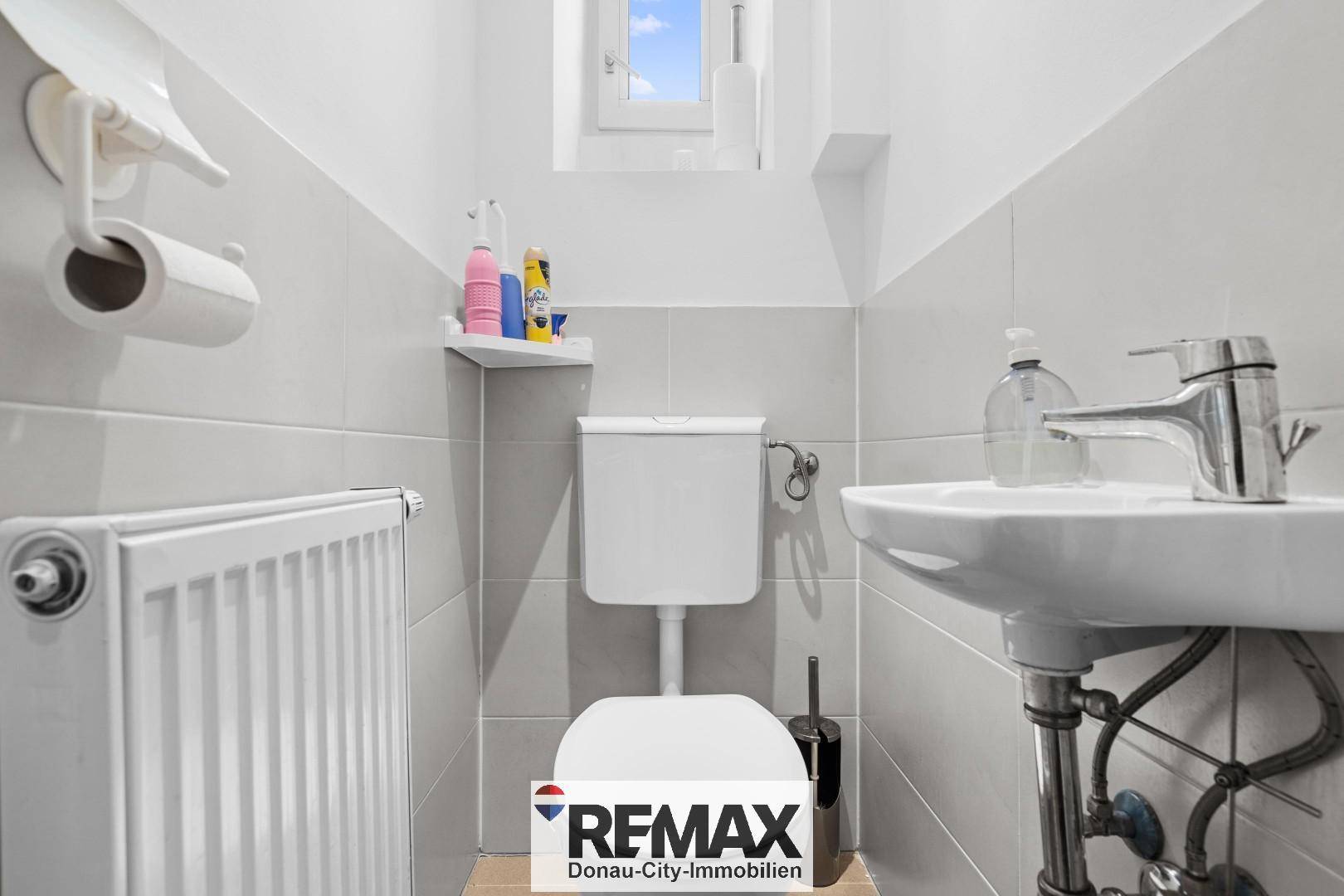 Separate Toilette mit Fenster, Waschbecken und Heizkörper, in hellen Fliesen gehalten.