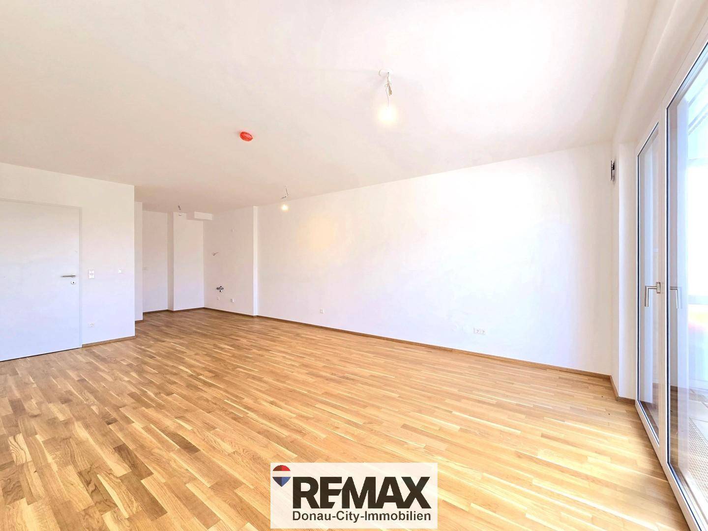 Geräumiges, leeres Wohnzimmer mit Holzboden und großen Fenstern, die viel Licht hereinlassen.