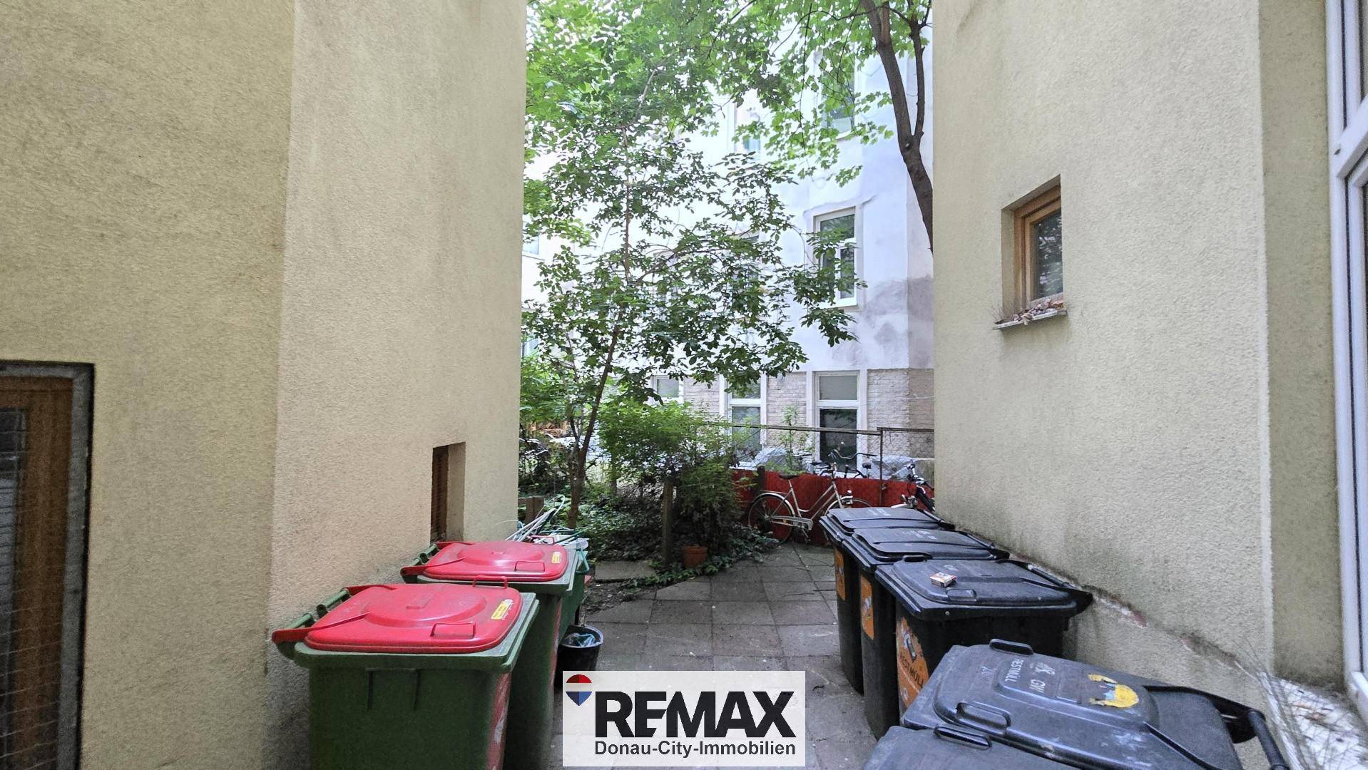 Hinterhofbereich mit Mülltonnen und Blick auf grüne Bäume im Innenhof eines Mehrfamilienhauses.