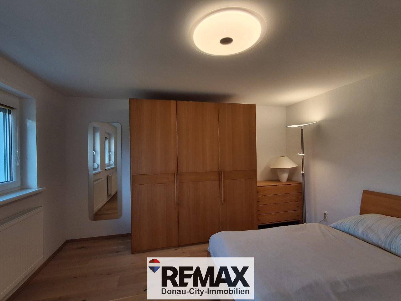 Schlafzimmer mit Holzkleiderschrank, Bett und Fenster, das viel Tageslicht hereinlässt.