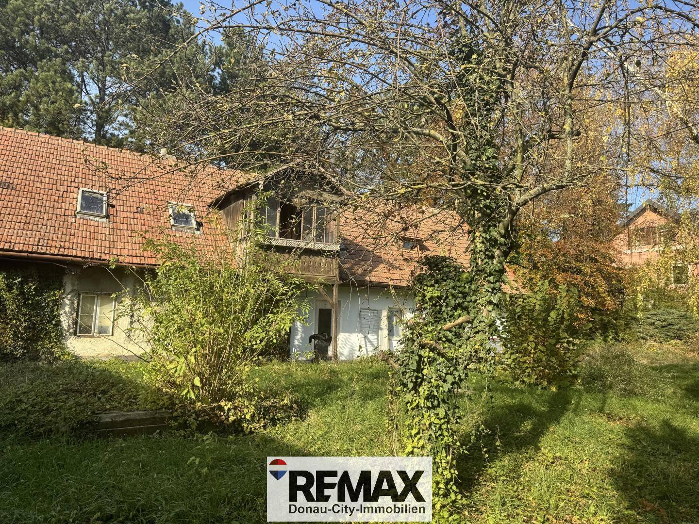 Ansicht des alten Hauses mit Holzbalkon und Ziegeldach, umgeben von einem großen, baumbestandenen Garten.