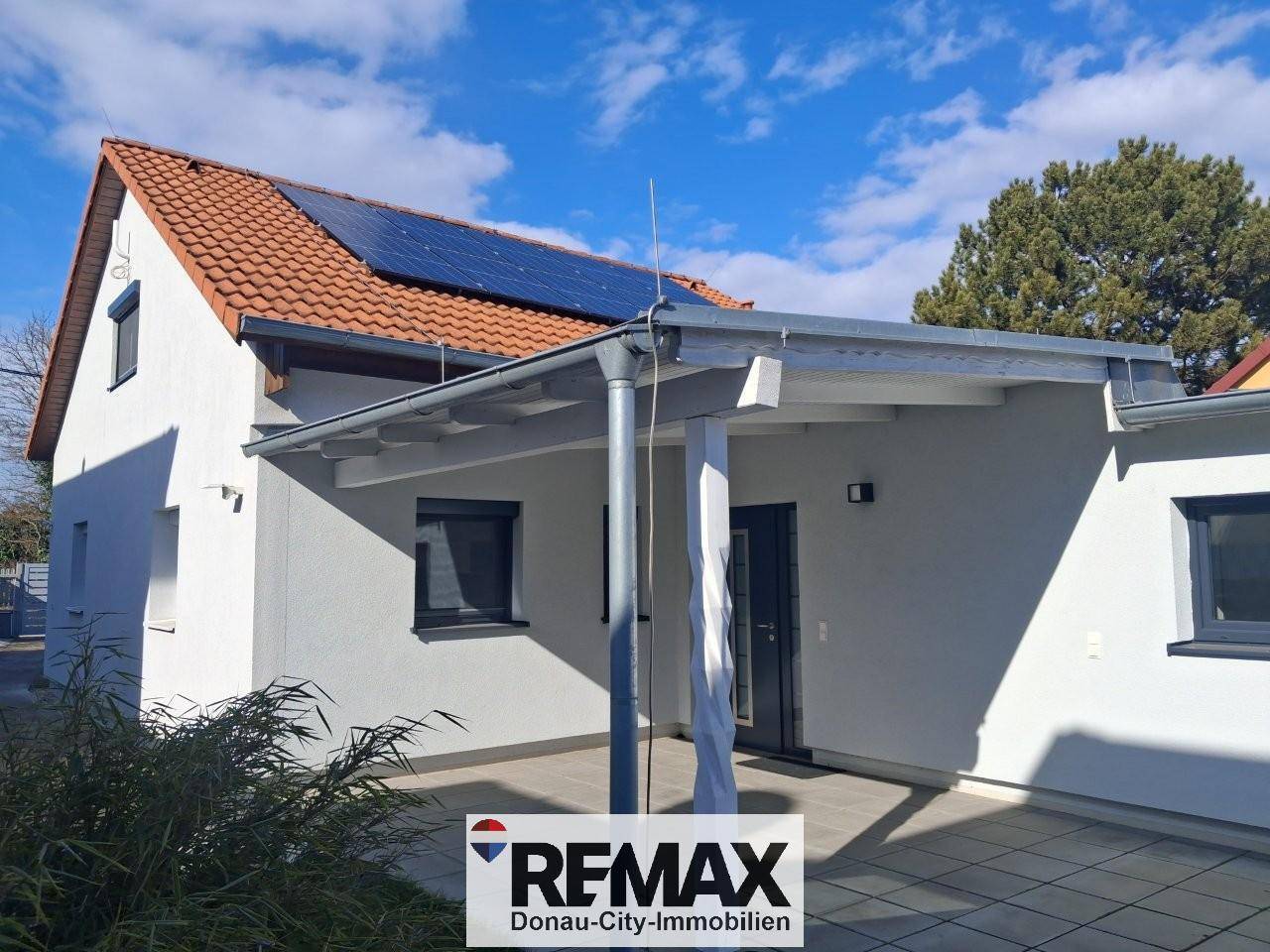 Hausfassade mit rotem Ziegeldach, Solaranlage und überdachter Terrasse im Außenbereich.