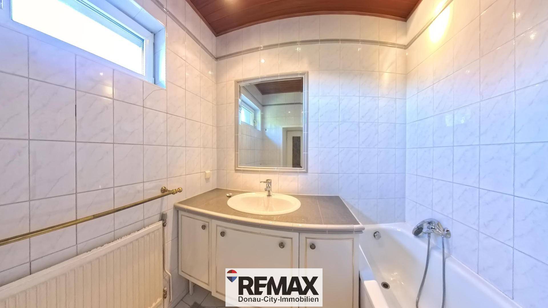 Badezimmer mit weißen Fliesen, Waschtisch und Badewanne, ausgestattet mit einem Fenster für Tageslicht.