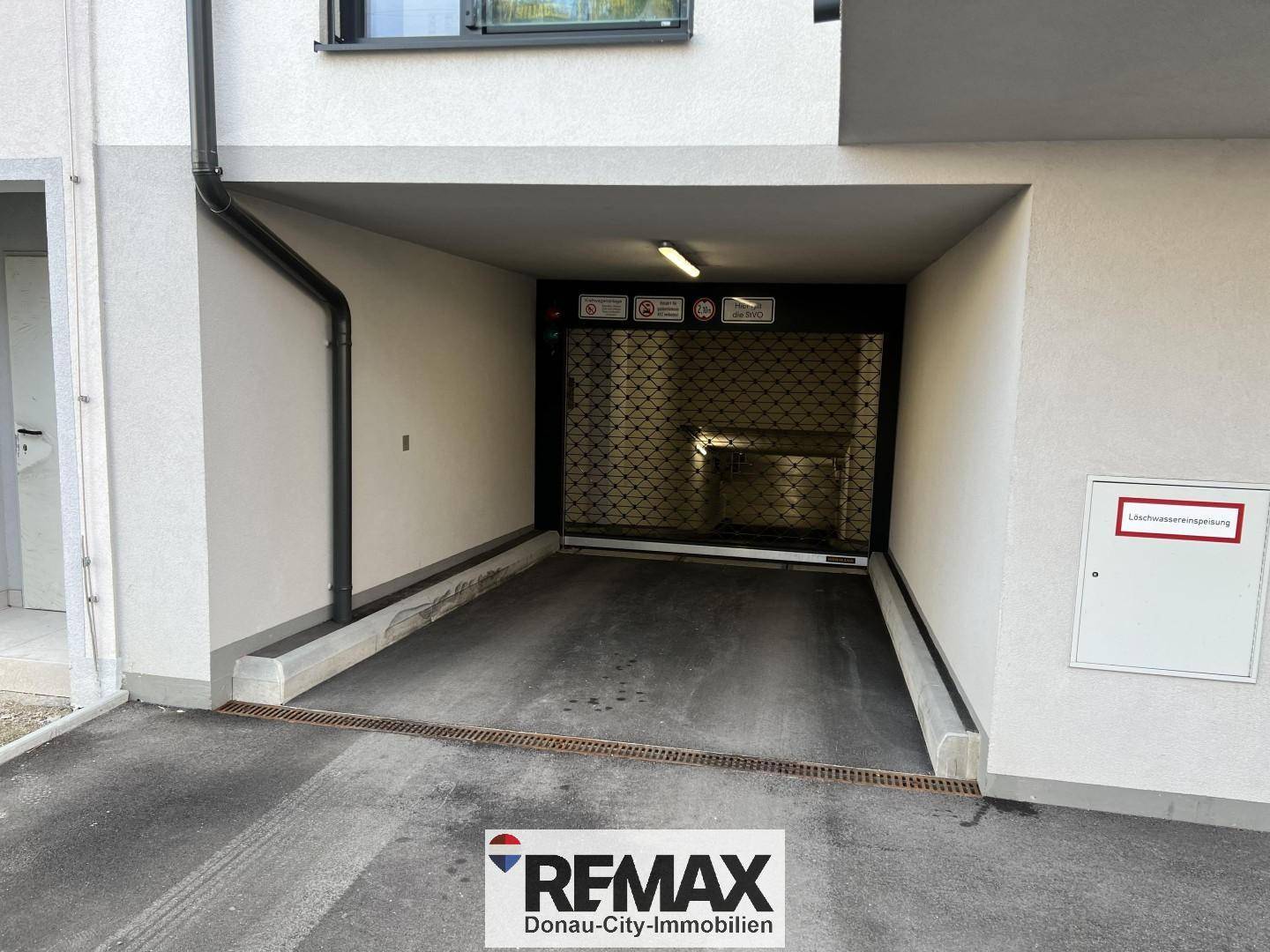 Eingang zu einer Tiefgarage mit Rolltor und Hinweisschildern an der Fassade eines Neubaus.