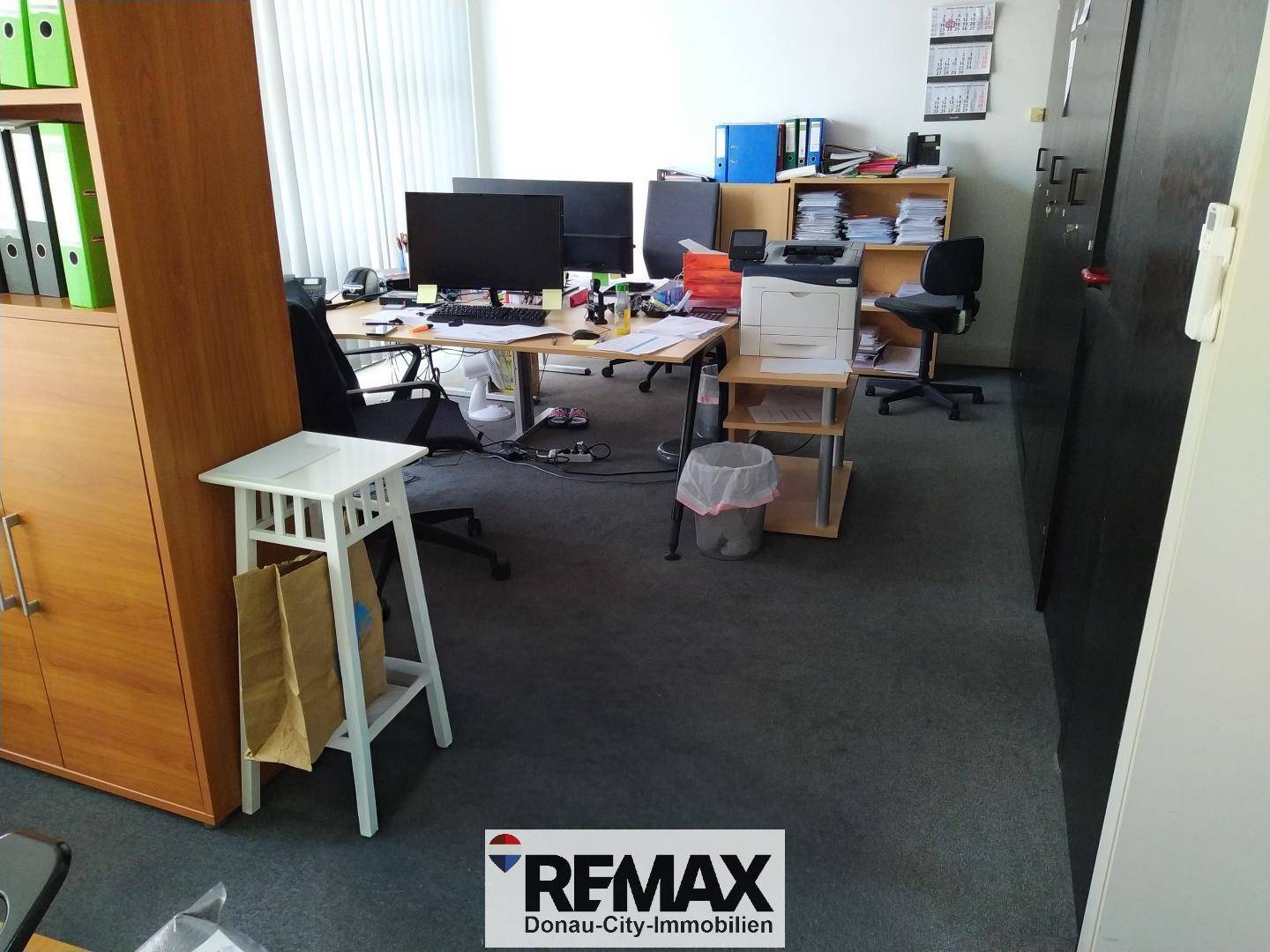 Büro mit mehreren Arbeitsplätzen, Teppichboden und vielen Aktenordnern in Regalen.