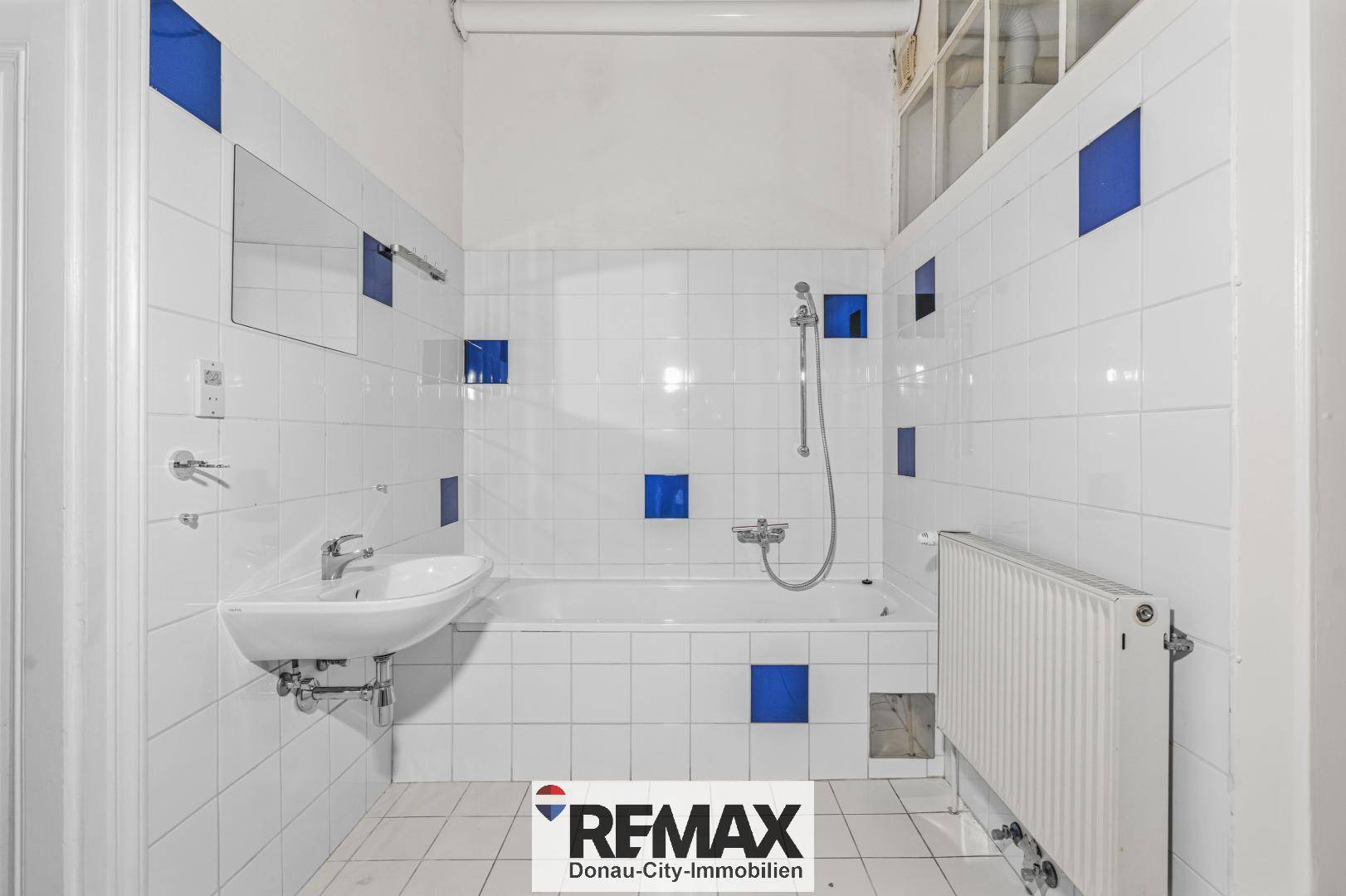 Badezimmer mit weißen Fliesen, blauen Akzenten, Badewanne mit Duschfunktion und einem Waschbecken.