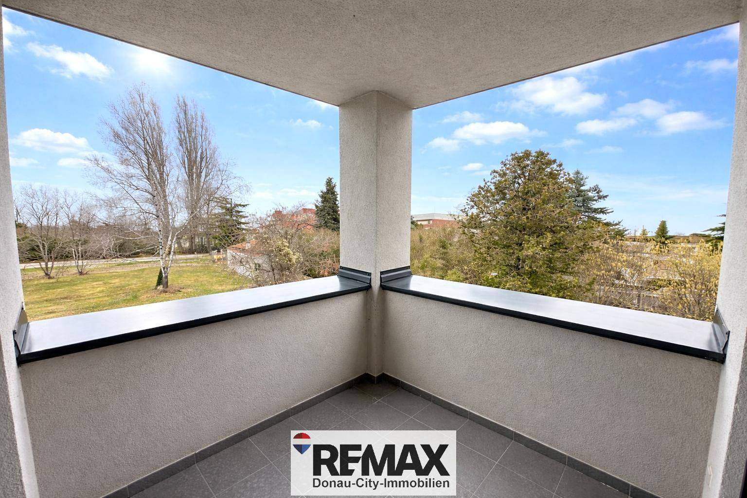 Heller Balkon mit Fliesenboden und weitem Blick ins Grüne mit Bäumen.