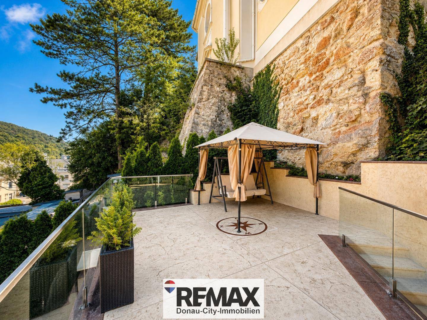 Sonniger Terrassenbereich mit Schaukel und Pavillon, umgeben von einer Steinmauer.