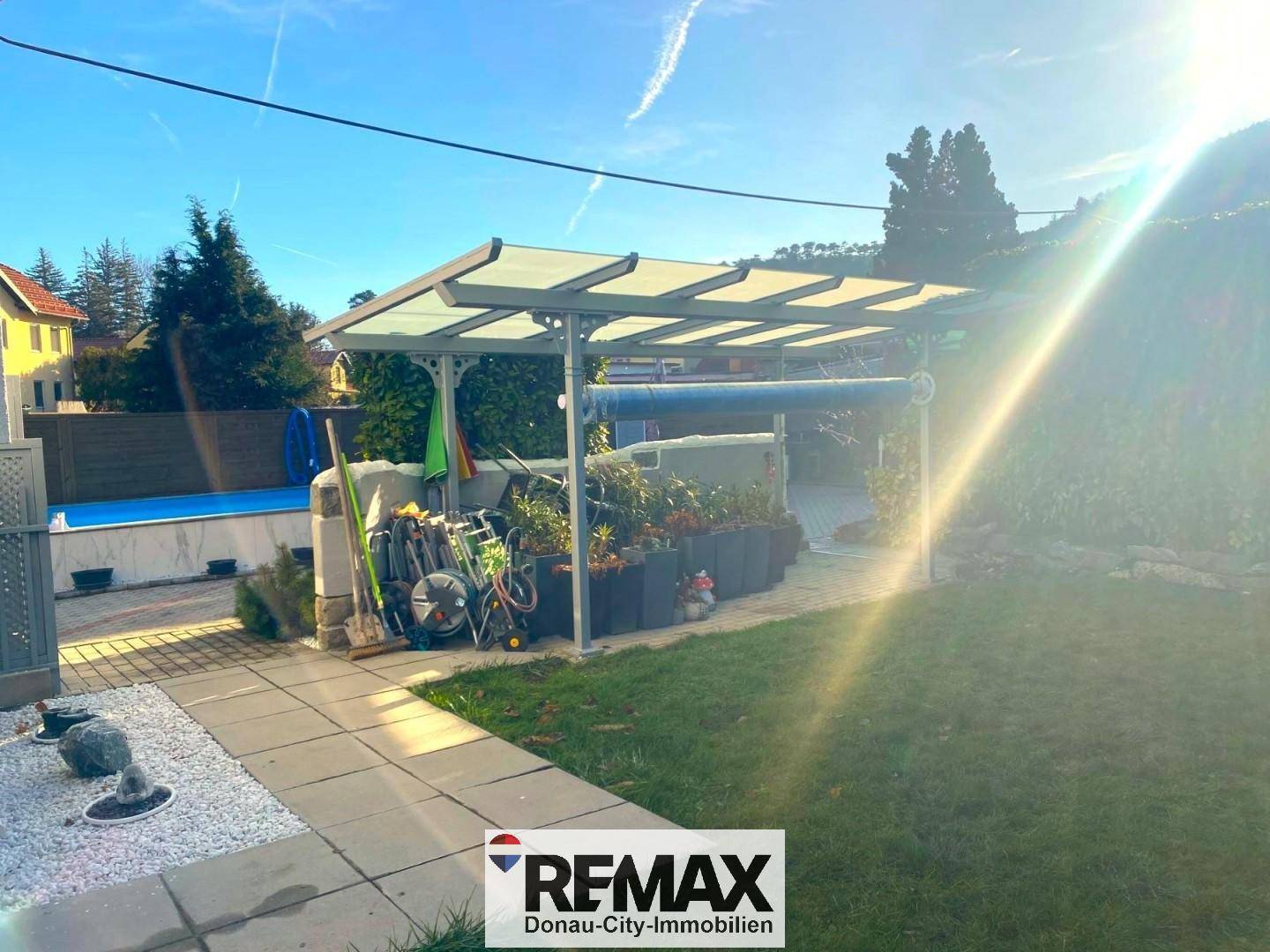 Sonniger Gartenbereich mit Carport und Rasenfläche, im Hintergrund ist der Pool sichtbar.