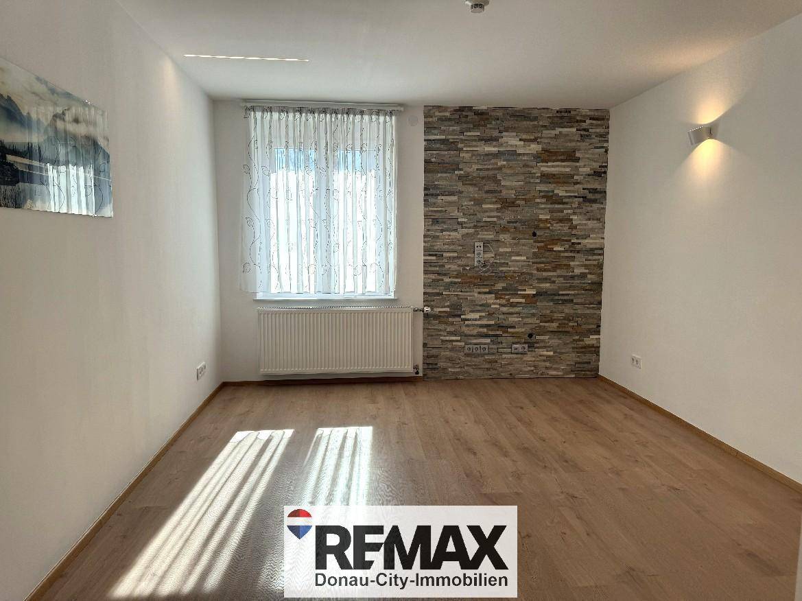 Leeres Wohnzimmer mit hellem Laminatboden, dekorativer Steinwand und großem Fenster.