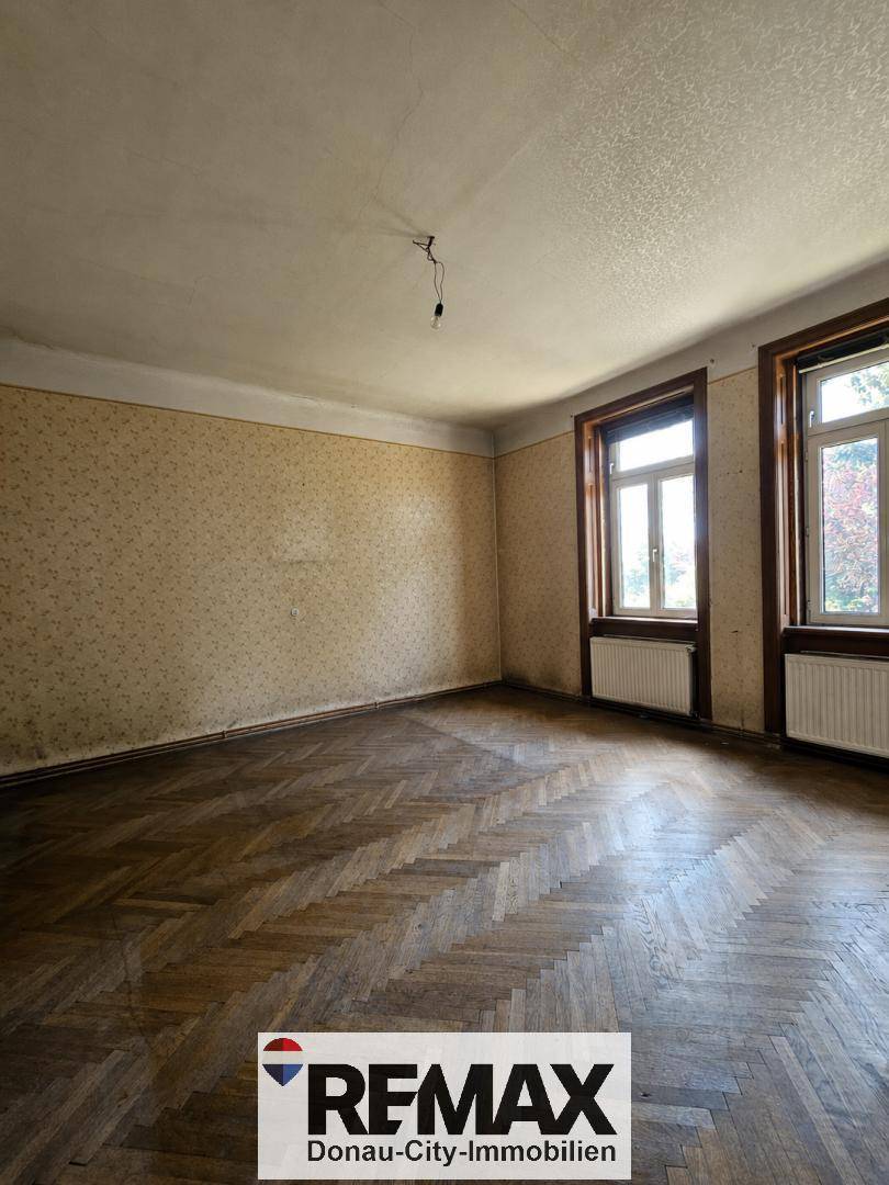 Leeres Wohnzimmer mit Fischgrätparkett, zwei Fenstern und alter Tapete, bereit zur Renovierung.