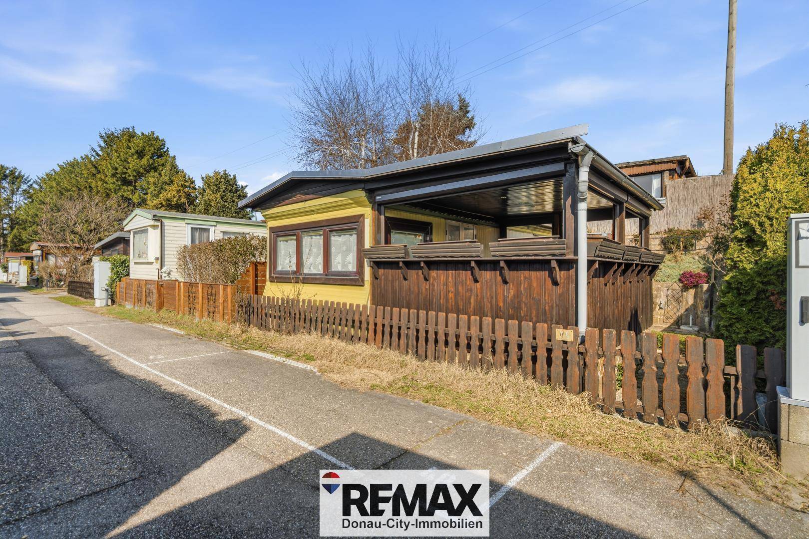 Kleines gelbes Haus mit überdachter Veranda und Holzzaun in einer Siedlung.