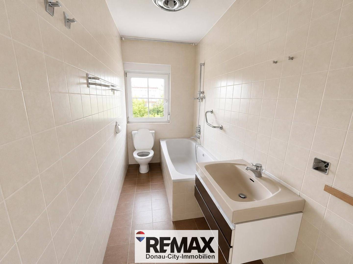 Badezimmer mit beigefarbenen Fliesen, Badewanne, WC, Waschtisch und einem Fenster für Tageslicht.