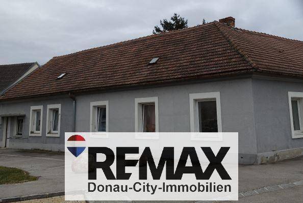 Vorderansicht des Hauses mit grauer Fassade, mehreren Fenstern und einem gepflasterten Bereich.